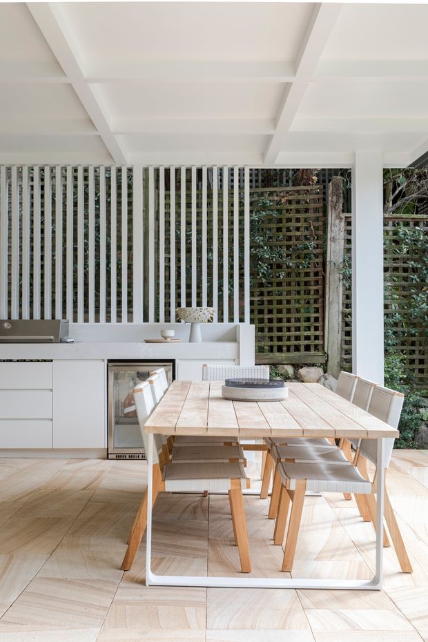 Bright covered outdoor dining and kitchen area with white structural beams, a fixed white kitchen run, timber dining table and chairs, pale timber flooring, and vertical and lattice screening opening onto greenery.