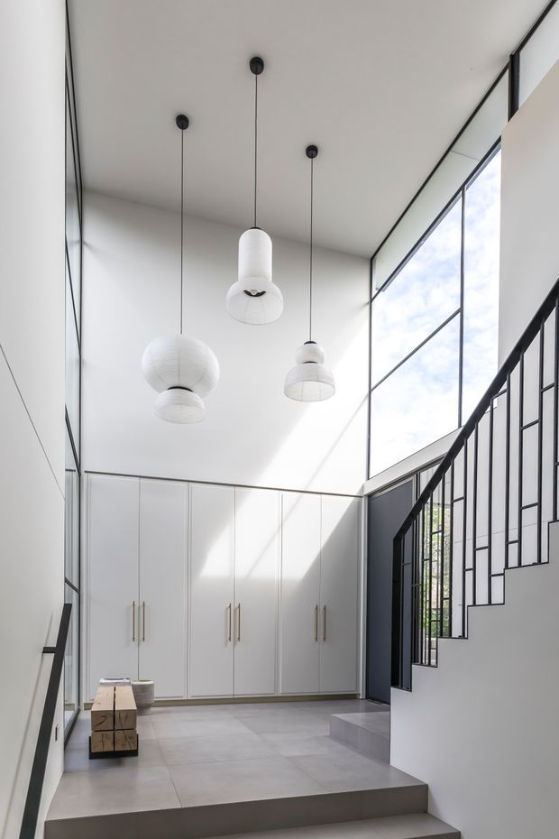 A bright, minimalist double-height entry hall with floor-to-ceiling glazing, white built-in cabinetry, pale tiled flooring, sculptural pendant lights, and a black metal staircase balustrade. Strong natural light fills the space, creating a clean, airy contemporary interior.