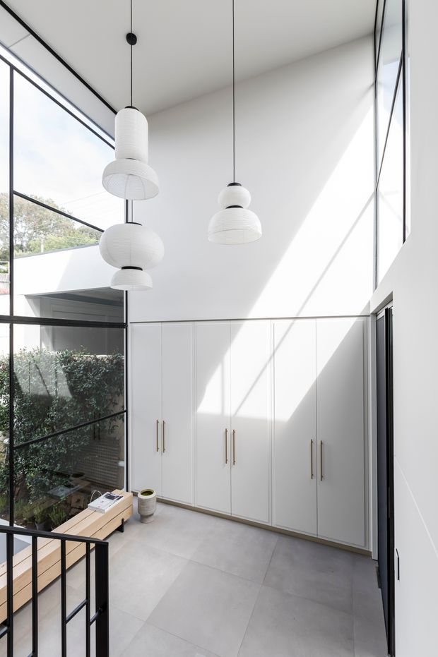 A bright, double-height interior with full-height black-framed glazing, white built-in cabinetry with long brass-toned handles, large pale grey floor tiles, and sculptural white pendant lights. A glazed internal courtyard or planted void is visible at left, with a stair or balustrade in the foreground. Strong sunlight creates geometric shadows across the walls and cabinetry.