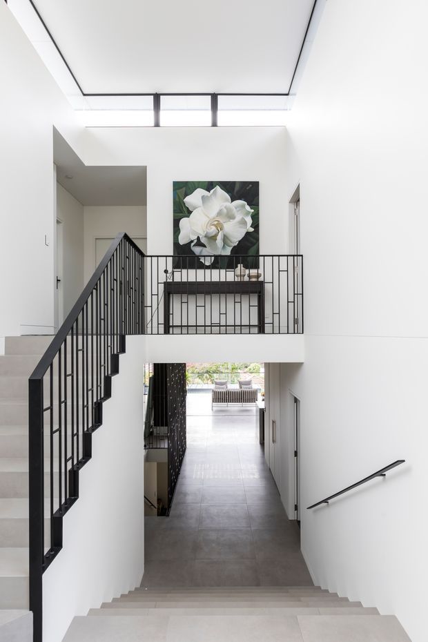 Bright contemporary interior view of a stairwell and landing. White walls and ceilings surround a two-level stair circulation space, with black metal balustrades and handrails, large grey floor tiles, a skylight or clerestory above, and a floral artwork over a console on the landing. The stairway continues down to a glazed outdoor area.