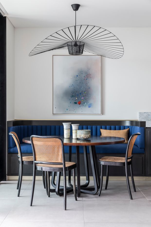 A refined dining area with a round dark dining table surrounded by woven-back dining chairs and a built-in deep blue banquette. Dark timber or painted joinery runs along the back wall beneath a large abstract artwork, while a sculptural black pendant light forms a prominent focal point overhead. The space has pale tiled flooring, white walls, and a sophisticated high-contrast palette.