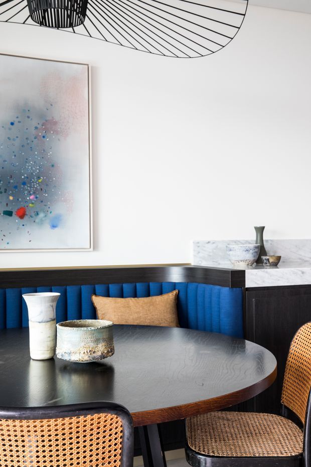 A refined dining nook with a round dark timber dining table, woven cane chairs, and a vivid deep-blue built-in banquette. A black timber or stained-wood cabinet runs along the wall, topped with a pale marble surface and decorative vessels. The white wall features a large abstract artwork, while a sculptural black pendant light hangs overhead. The scheme combines bold blue upholstery, dark joinery and timber, woven natural textures, and pale stone.