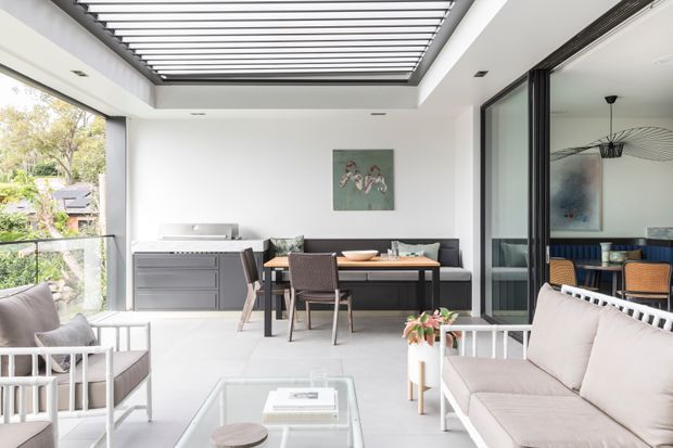 Bright contemporary open-plan living and dining area opening onto a covered outdoor entertaining space. The room features white walls and flooring, extensive black-framed glazing and sliding doors, a linear skylight or louvred roof opening, a dark dining table with upholstered chairs, a dark sofa, pale lounge seating, and a built-in outdoor-style kitchen or barbecue along the rear wall. A secondary dining area is visible through the glazing.
