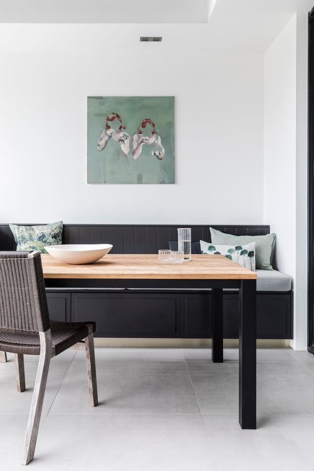 Bright contemporary dining nook with a built-in black banquette along the wall, a light timber dining table, woven dining chairs, and pale grey tiled flooring. White walls and ceiling create an airy backdrop, with botanical cushions and abstract artwork adding muted green accents.