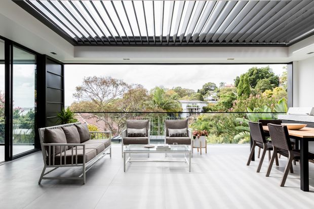 Bright contemporary open-plan living and dining area opening onto a balcony or terrace, with floor-to-ceiling glazing, a large retractable louvred roof, neutral tiled flooring, black-framed sliding doors, and views over a lush suburban landscape. The space includes an outdoor-style lounge setting and a timber dining table.