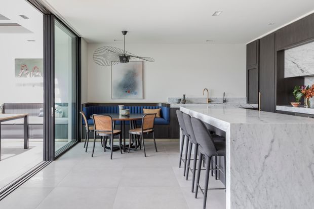 A bright, contemporary open-plan kitchen and dining area with a large white marble island, grey upholstered bar stools, dark timber cabinetry, and a built-in navy banquette. A small dining table with woven chairs sits beside full-height glazed sliding doors opening to an outdoor area. The space features light stone-look tiled flooring, white walls, a sculptural pendant light, and abundant natural light.