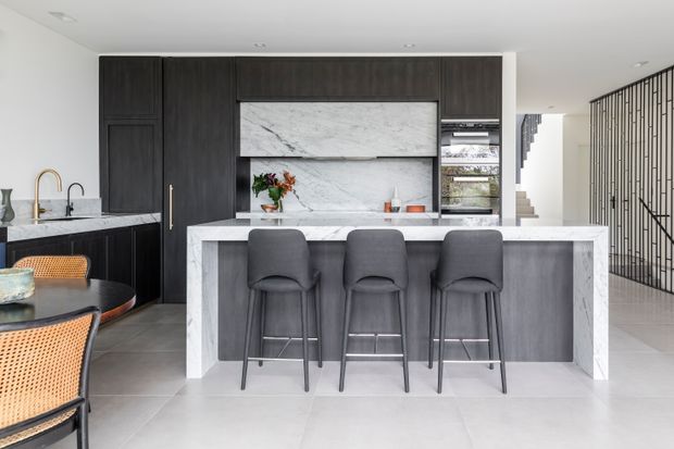 Contemporary open-plan kitchen with a large waterfall-edge island and three bar stools, dark timber cabinetry, pale veined stone benchtops and splashback, integrated appliances, and a black metal stair balustrade visible beyond. A dining area is partially visible in the foreground.