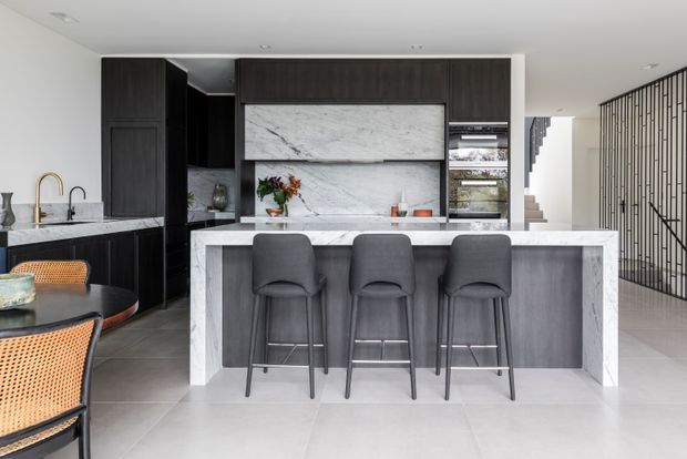 Contemporary open-plan kitchen dominated by a large marble-clad island with three dark upholstered bar stools. The space features dark timber cabinetry, a marble splashback and benchtop, integrated appliances, pale large-format flooring, and a black vertical balustrade beside the staircase. A dining area is partially visible at left.