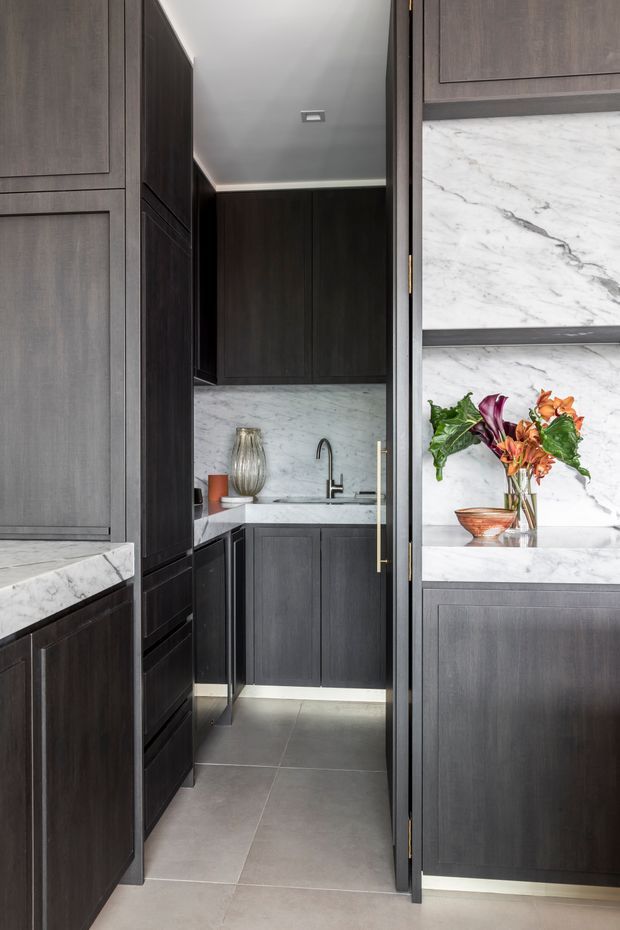 A compact, refined kitchen or butler’s pantry with floor-to-ceiling dark timber cabinetry, white veined marble benchtops and splashback, a stainless or dark metal mixer tap, and large grey porcelain floor tiles. Brass-toned cabinet hardware and concealed under-cabinet lighting add warmth and polish. A sink and preparation area are visible at the rear, framed by tall storage cabinetry.
