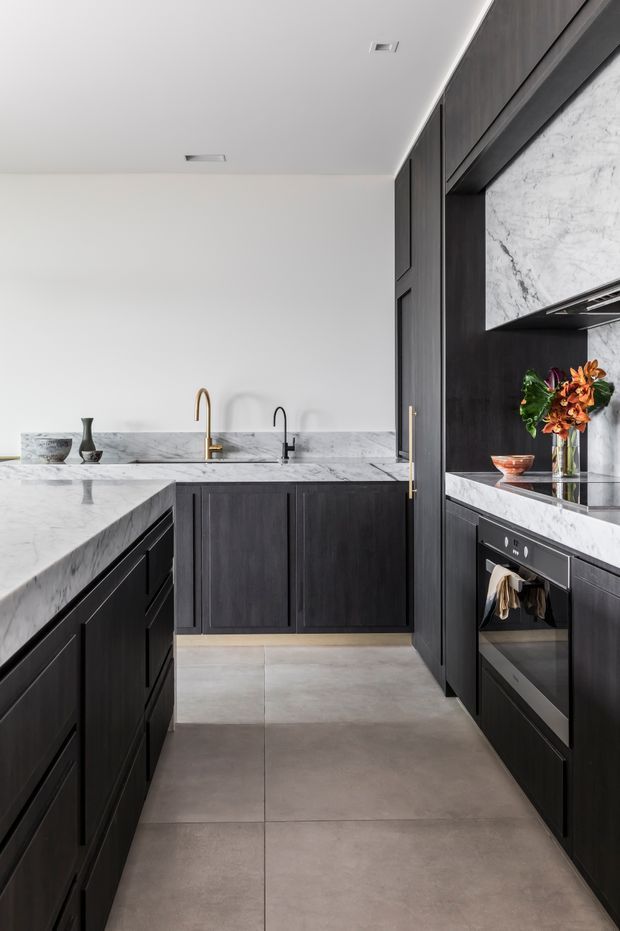 A refined modern kitchen with a galley-style layout and dark timber cabinetry. Large white-grey veined stone surfaces form the island, benchtops and splashback, complemented by integrated appliances, a sculptural rangehood surround, and contrasting brass and black tapware. Large-format grey floor tiles and recessed ceiling lighting complete the clean, high-contrast scheme.