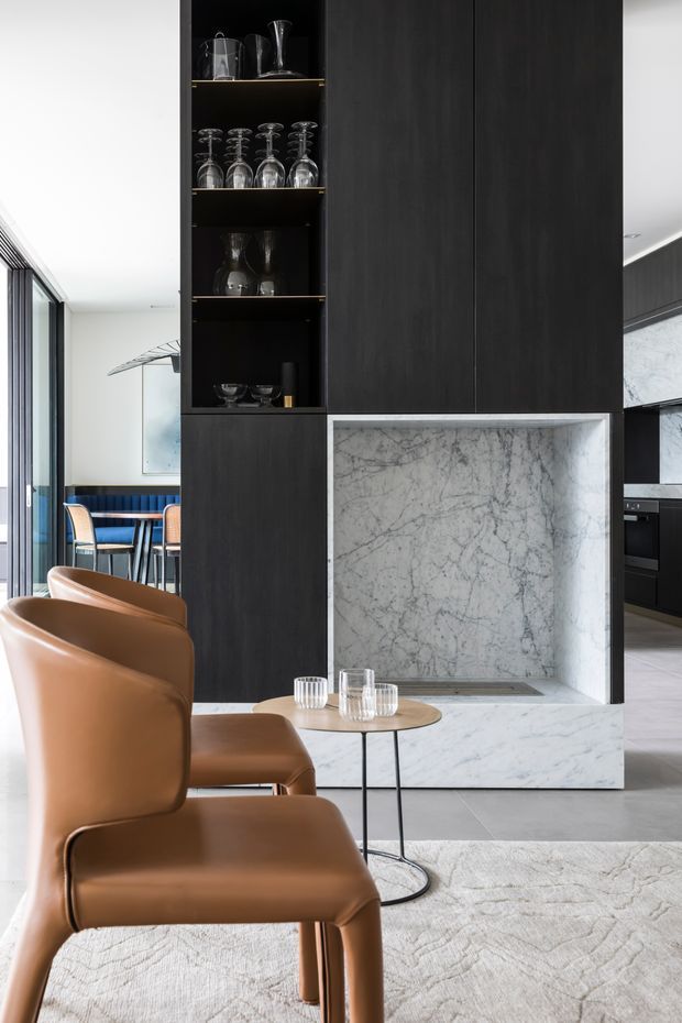 Contemporary open-plan living and dining area featuring a dramatic black timber-look built-in cabinet and open glassware shelving, a marble fireplace surround with recessed hearth, caramel leather dining chairs, a small round side table, and pale stone flooring with a textured light rug. Floor-to-ceiling glazing and a blue-upholstered dining area are visible to the left.