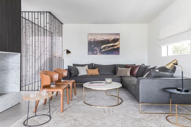 Bright contemporary living room with a large dark grey sectional sofa, leather dining-style armchairs, round glass coffee table, marble fireplace surround, patterned rug, and sculptural floor and table lamps. A slim black metal screen creates a stairway partition at the rear, while a small window and white Roman blind add natural light.