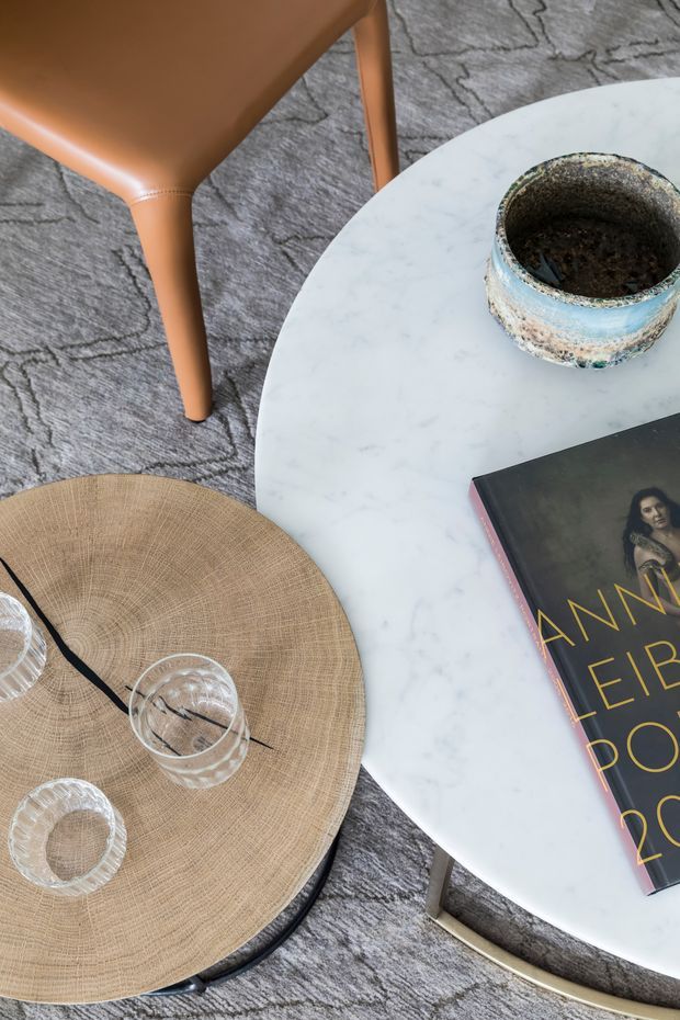 Close-up interior vignette showing a round white marble coffee table with a weathered blue ceramic vessel and a fashion book, paired with a round woven ottoman or tray and clear glassware. A tan leather chair and grey patterned rug complete the refined contemporary lounge setting.