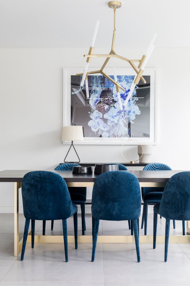 A bright, refined dining room featuring a long rectangular dining table with a pale stone or light-toned top and gold-toned base, surrounded by deep blue velvet upholstered dining chairs. A sculptural brass-gold pendant light hangs above the table, while a framed abstract artwork, table lamp, and small decorative objects add visual interest. The room has white walls and large-format pale flooring tiles.