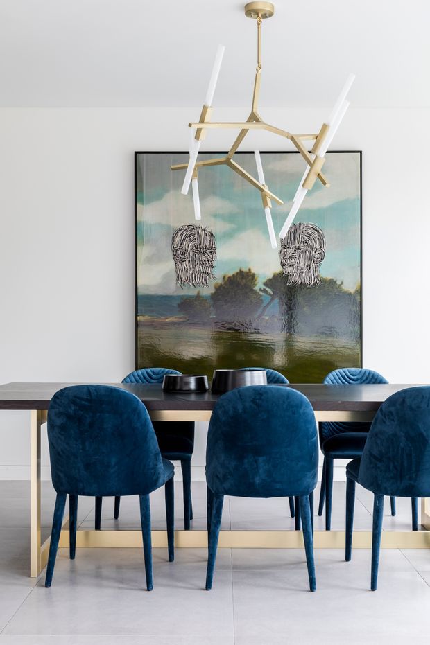 A contemporary dining room with a long dark dining table surrounded by deep blue upholstered chairs. A sculptural brass-and-white pendant light hangs above the table, while a large framed landscape artwork with abstract portrait figures forms a dramatic focal point against the pale wall. The space has a bright, refined palette with light tiled flooring and minimal architectural detailing.
