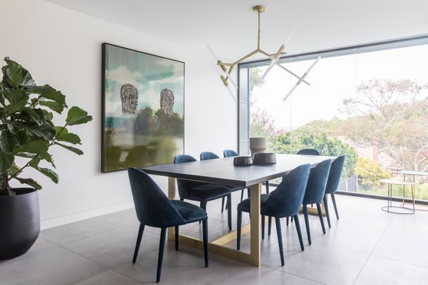 Bright contemporary dining room with a long dark dining table, blue upholstered dining chairs, a sculptural brass-and-white pendant light, large abstract artwork, a potted indoor plant, and expansive corner glazing overlooking greenery.