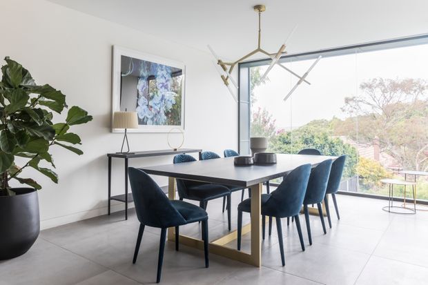 Bright contemporary dining room with a long dark dining table, upholstered navy-blue dining chairs, a slim console table, framed artwork, a large potted plant, and sculptural pendant lighting. Floor-to-ceiling glazing opens to leafy outdoor views, creating an airy, refined setting.