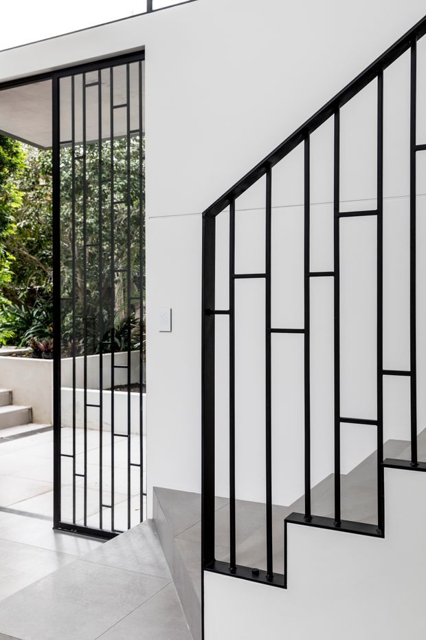 A minimalist interior stairway with a geometric black metal balustrade against crisp white walls. Large black-framed glazed doors open toward a planted courtyard, while pale grey floor tiles and concrete stair surfaces create a clean, contemporary composition.