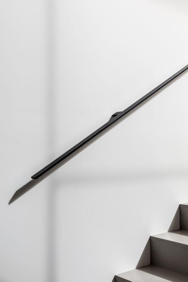 Minimalist architectural detail showing a slim black metal stair handrail mounted diagonally on a white plaster wall beside a modern grey stair flight. Strong directional sunlight casts a sharp shadow, emphasizing the clean geometric composition.