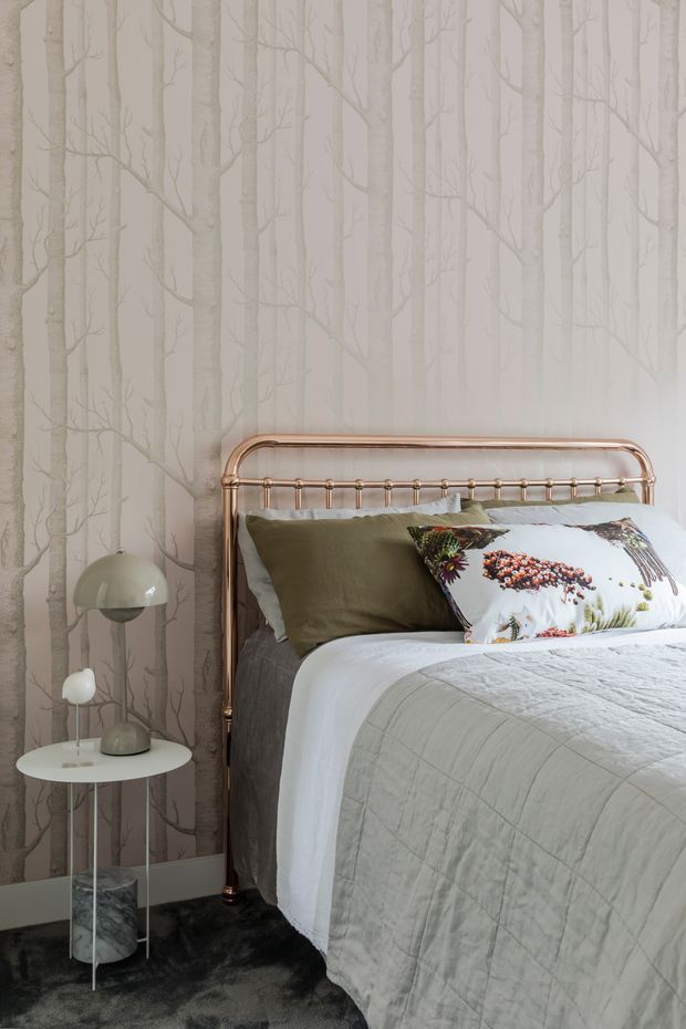 A softly styled bedroom featuring a copper-toned metal bed frame, layered grey and olive bedding, and a botanical-patterned accent pillow. The walls are covered in pale tree-pattern wallpaper, complemented by a small round white side table, sculptural table lamps, and a dark plush carpet.