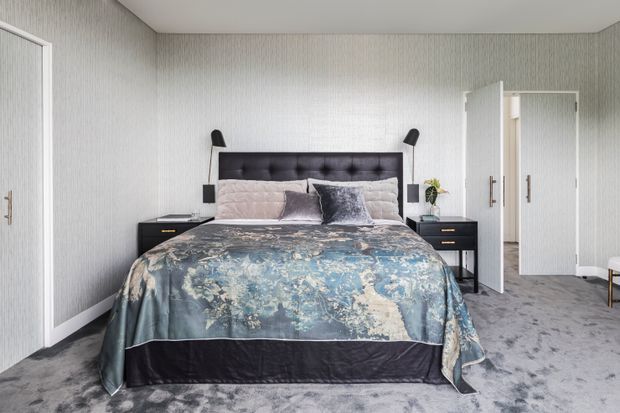 A contemporary bedroom with a large upholstered, button-tufted dark headboard, bedside tables, wall-mounted black reading lights, layered pale bedding, and a blue-grey floral bedspread. The room has pale vertically textured wallcovering, grey carpet, white trim, and built-in wardrobes with open doors.