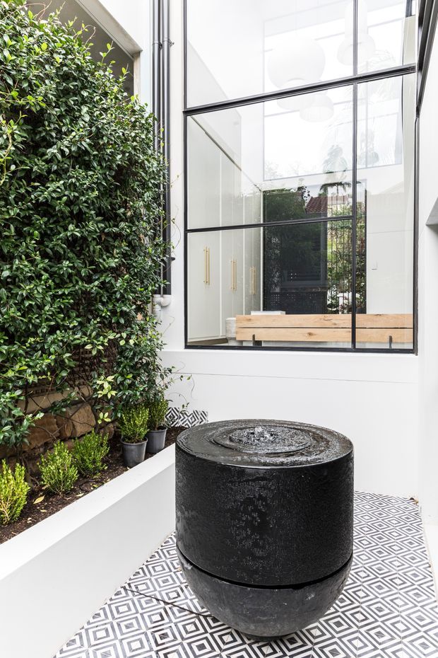A bright, refined courtyard or outdoor area with a black cylindrical water feature positioned on patterned geometric floor tiles. Dense green planting lines one side, while white walls and large black-framed glazed openings create a contemporary indoor-outdoor connection. A timber bench and pale cabinetry are visible through the glazing.