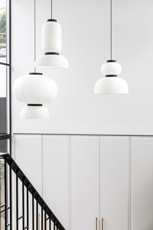Bright minimalist interior view of a stairwell or landing, featuring a tall white paneled wall with integrated cabinetry, black metal stair balustrade, large black-framed glazing at left, and a cluster of sculptural white ribbed pendant lights suspended at different heights.