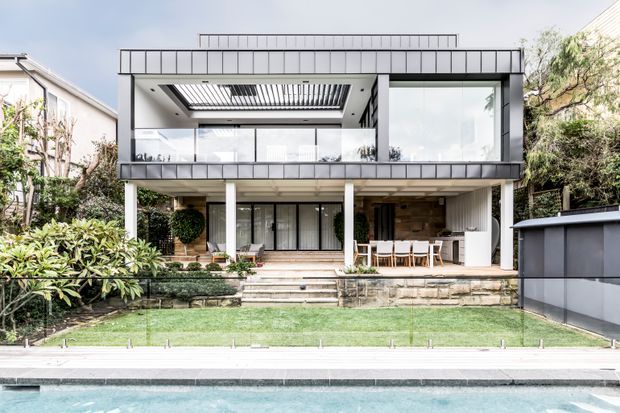 A contemporary two-storey residential house viewed from the rear garden, with broad floor-to-ceiling glazing, a strongly horizontal upper-level form, a covered ground-floor outdoor dining area, landscaped lawn, and a swimming pool in the foreground. The facade combines dark metal-framed glazing, pale rendered or painted surfaces, and timber-look wall cladding beneath a flat roof.