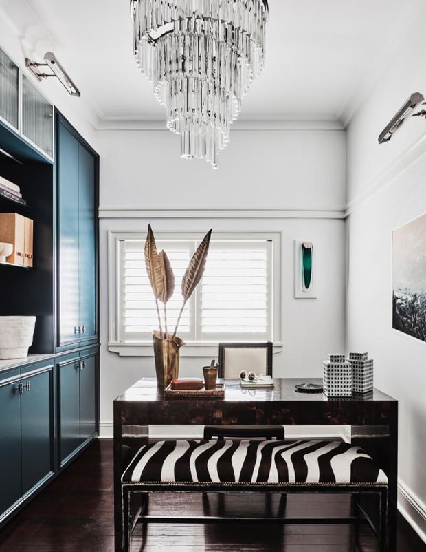 A refined home office with a compact dark wood desk, black-and-white striped bench, deep teal built-in cabinetry, white plantation shutters, and a dramatic tiered crystal chandelier. Classic wall mouldings, polished dark flooring, and carefully styled accessories create a high-contrast, elegant interior.