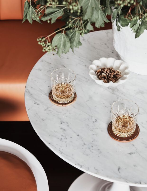 Close-up styled dining setting featuring a round white-grey veined marble table, two cut-crystal tumblers on brown woven coasters, a white scalloped bowl of walnuts, and a large white planter with leafy greenery. Curved white dining chairs with rust-orange upholstery surround the table, creating a refined, warm, contemporary atmosphere.
