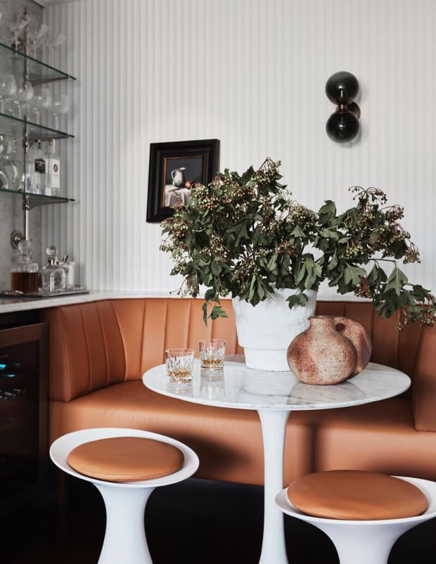 A sophisticated dining nook with a curved cognac-brown upholstered banquette, white pedestal stools, and a round white marble table. Vertical white wall panelling, black glass shelving stocked with glassware, a dark double-sphere wall light, framed artwork, and botanical arrangements create a refined, moody setting.