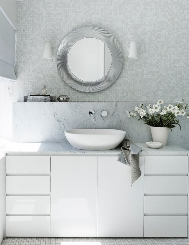 Bright, refined contemporary bathroom featuring a white floating vanity with integrated drawer cabinetry, a veined marble benchtop and backsplash, a white vessel basin, round fluted-framed mirror, pale mosaic wall tiles, and minimalist wall sconces. Soft grey textiles and a potted floral arrangement add subtle warmth.
