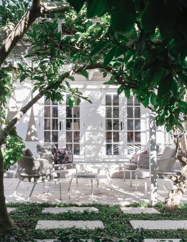 A bright, elegant courtyard patio framed by mature leafy trees, with white French doors and tall multi-pane windows opening onto a light paved terrace. The outdoor seating arrangement includes woven lounge chairs, a sofa, a low round coffee table, and neutral cushions, with stepping stones through a planted groundcover garden in the foreground. The scene has a refined, classic coastal character and strong dappled sunlight.