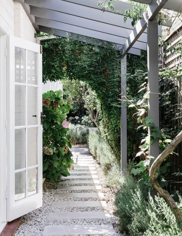 A narrow, lush garden walkway viewed from beneath a grey timber pergola. A white glazed door opens onto the path at left, while stepping slabs set in pale gravel lead through dense greenery, hydrangeas, shrubs and climbing plants. Timber screening and pergola supports define the enclosed outdoor passage.
