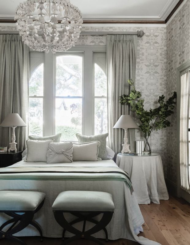 Elegant traditional bedroom with a pale upholstered bed, layered white and grey bedding, matching stools, tall arched windows with full curtains, ornate floral wallpaper, decorative moulding, crystal chandelier, bedside lamps, and a round draped side table. The room has a refined, softly lit, monochromatic palette with warm timber flooring.