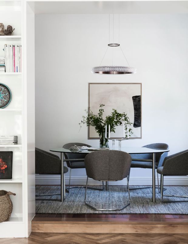 A refined dining area viewed through a wide opening, featuring a dark oval dining table with upholstered chairs, a contemporary suspended pendant light, framed artwork, and a vase of greenery. White built-in shelving lines the left side, while a textured rug sits over a herringbone timber floor.