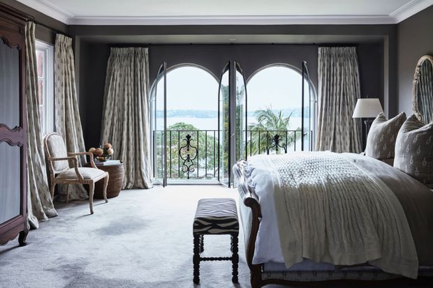 Elegant bedroom with dark charcoal walls, ornate white crown moulding, full carpeting, patterned drapery, and a large upholstered bed. Tall arched glazed doors open to a balcony with decorative wrought-iron railing and views beyond. A carved wardrobe, wicker side table, armchair, bedside lamp, and upholstered bench complete the refined interior.