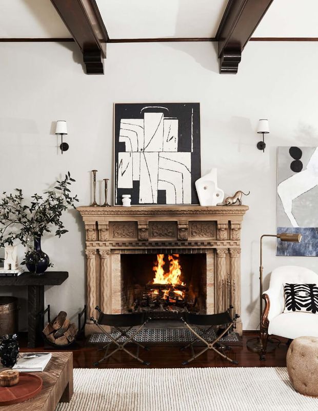 A refined living room centred on an ornate carved stone fireplace with a lit fire, dark timber ceiling beams, and a curated arrangement of contemporary and traditional furnishings. Abstract artwork, sculptural decor, wall sconces, a leather-and-metal firewood bench, armchair, coffee table, and neutral rug create a warm, eclectic setting.