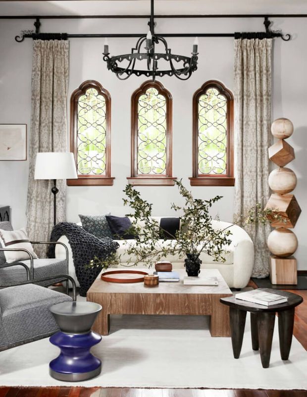 A refined living room with three tall arched stained-glass windows in timber surrounds, patterned curtains, a black wrought-iron chandelier, and a light sofa arranged around a large timber coffee table. Layered lounge chairs, sculptural side tables, botanical branches, artwork, and a neutral rug create an eclectic, sophisticated interior.