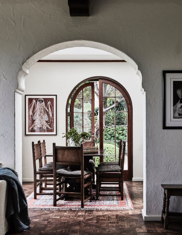 Interior view of a compact dining area framed by a sculpted arched opening. A dark timber dining table and chairs sit on a patterned rug, with a distinctive arched timber-framed glazed door opening onto greenery. Textured grey plaster walls, dark parquet flooring, framed artwork, and traditional timber detailing create a warm, characterful atmosphere.