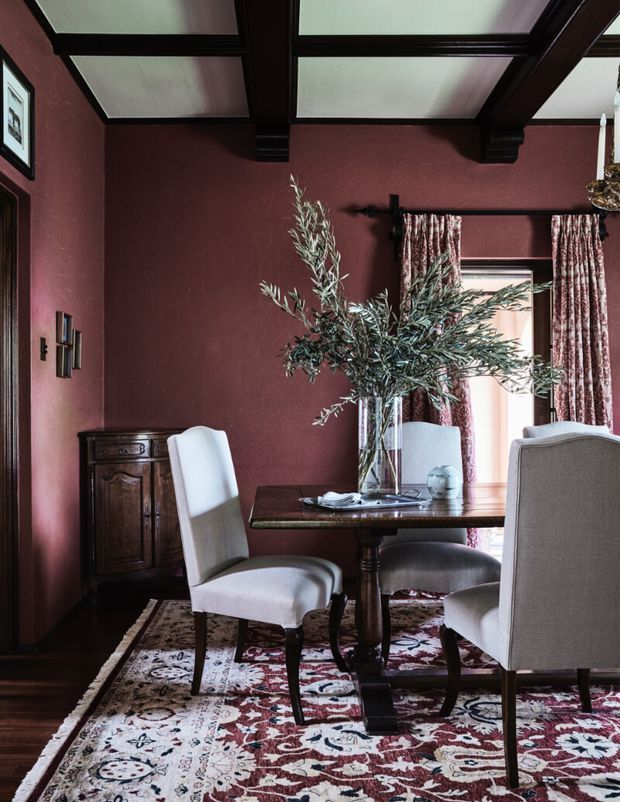 A richly styled formal dining room with deep burgundy walls, dark exposed ceiling beams, a traditional wooden dining table, upholstered dining chairs, patterned curtains, and a large Persian-style rug. An antique wooden sideboard and framed artwork add classic character, while a tall leafy arrangement forms the table centrepiece. The room is moody, warm, and refined.