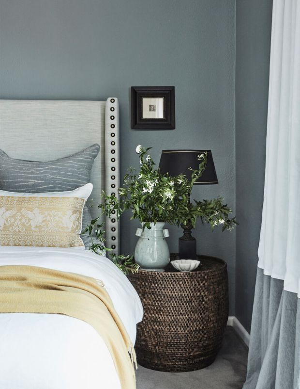 A refined bedroom corner featuring a partially visible upholstered bed with nailhead trim, layered white and patterned bedding, a woven round bedside table, decorative lamp, ceramic vase with greenery, framed artwork, and floor-length curtains. The room uses a muted blue-grey wall, soft neutrals, and warm yellow accents.