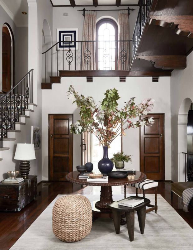A grand double-height residential foyer or living hall with a dark timber staircase and mezzanine gallery, ornate black metal balustrades, arched openings, tall arched window, white plaster walls, and dark timber floors. A round central table with decorative branches, upholstered stools, a woven ottoman, console furniture, and layered neutral furnishings create a refined traditional interior.
