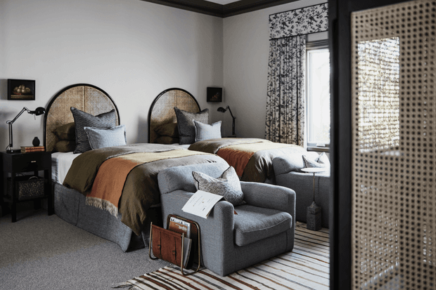 A refined twin bedroom with two upholstered beds positioned side by side, each featuring a rounded woven rattan headboard and layered neutral bedding with muted rust and olive accents. A grey lounge chair sits at the foot of the beds beside a small magazine rack, while dark bedside tables, black reading lamps, patterned window treatments, and a large woven-panel sliding door frame the room. The palette is warm, neutral, and textural with a calm, boutique-hotel atmosphere.