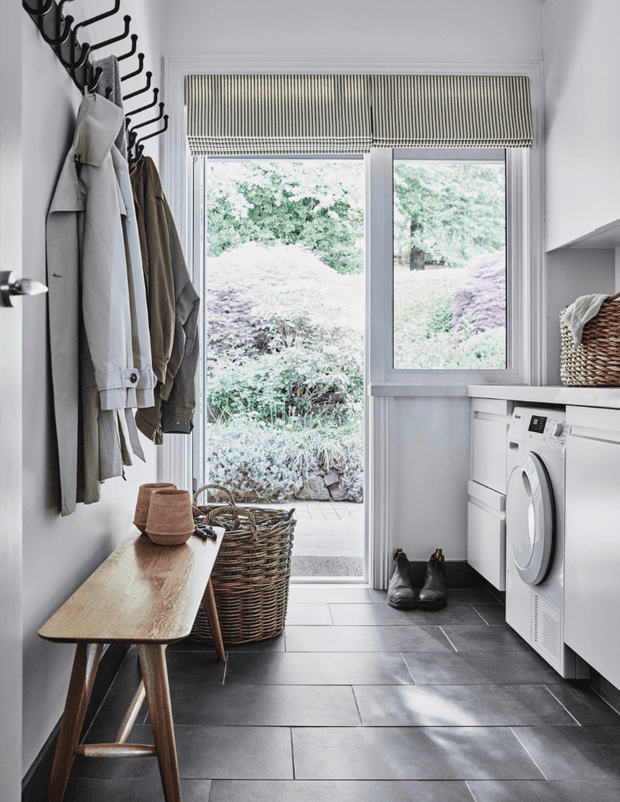 Bright, narrow laundry and mudroom with dark large-format tiled flooring, white cabinetry and a front-loading washer and dryer beneath a white countertop. A wall-mounted row of black hooks holds coats beside a simple timber bench, woven basket, and boots. A glazed door and window open to a landscaped garden, with woven roller blinds filtering daylight.