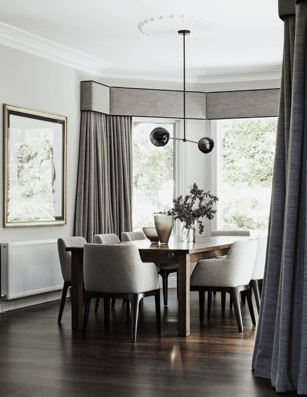 Elegant formal dining room with a dark timber dining table and upholstered dining chairs arranged beneath a sculptural pendant light. Tall bay windows are dressed with full-height grey curtains and box pelmets, while white walls, decorative ceiling mouldings, dark timber flooring, and framed artwork create a refined, tailored interior.