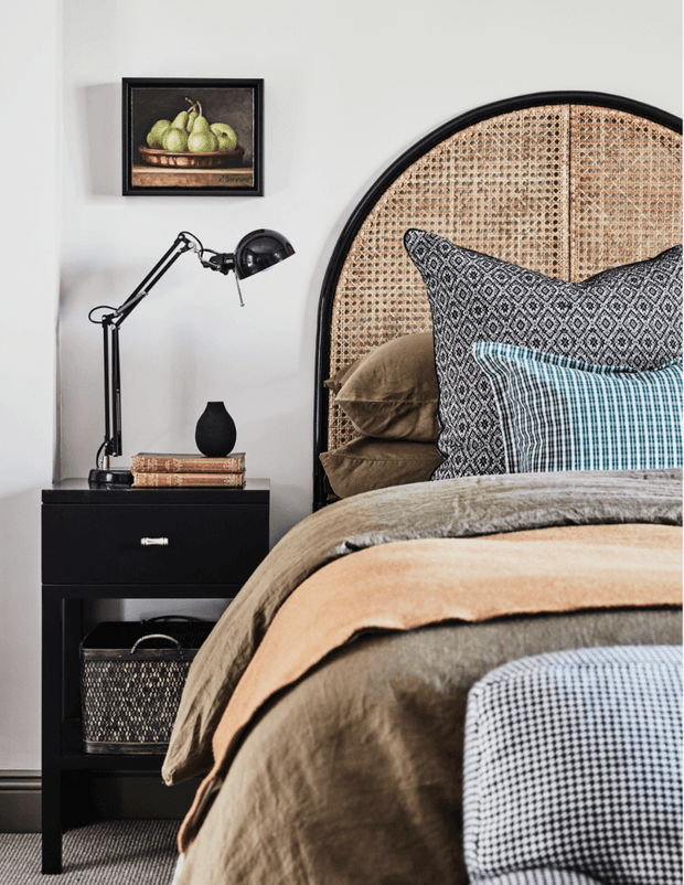 A styled bedroom vignette featuring a black bedside table with an adjustable black lamp, stacked books, and a small black vase. The bed has layered brown, tan, patterned black-and-white, teal, and checked textiles, with a large arched rattan cane headboard in a black frame. A framed still-life artwork hangs on the light wall above the bedside table. The room has a warm, relaxed, eclectic feel.
