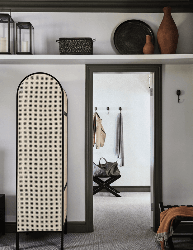 A refined interior vignette featuring dark charcoal trim, white walls, open display shelving, and carefully arranged decorative objects. A tall arched cane-panel screen stands in the foreground beside a doorway opening to a carpeted dressing or entry area with wall hooks, hanging textiles, and a folding bench. The palette is neutral and warm, with layered texture and traditional detailing.