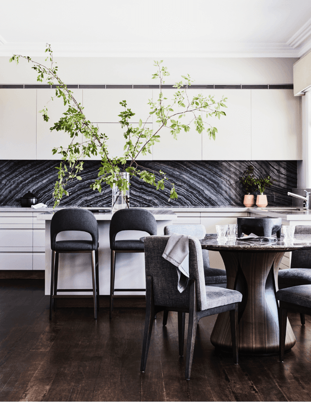 A refined open-plan kitchen and dining area with white built-in cabinetry, a dramatic black-and-white veined stone splashback, dark timber flooring, and a sculptural round dining table with upholstered chairs. A large branch arrangement adds an organic focal point, while layered ceiling mouldings and discreet linear ventilation contribute to the polished interior.