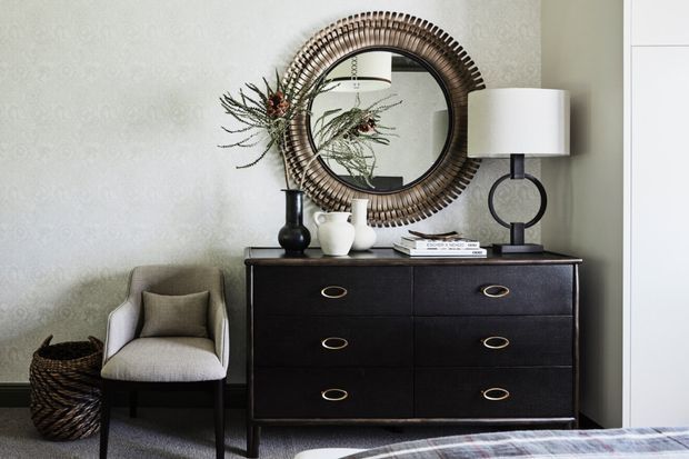 A refined bedroom vignette featuring a dark timber six-drawer dresser with brass pulls, a round woven-framed mirror, sculptural table lamp, decorative ceramics, and a light upholstered accent chair. The room has textured pale walls, neutral textiles, and layered natural materials, creating a warm, sophisticated atmosphere.
